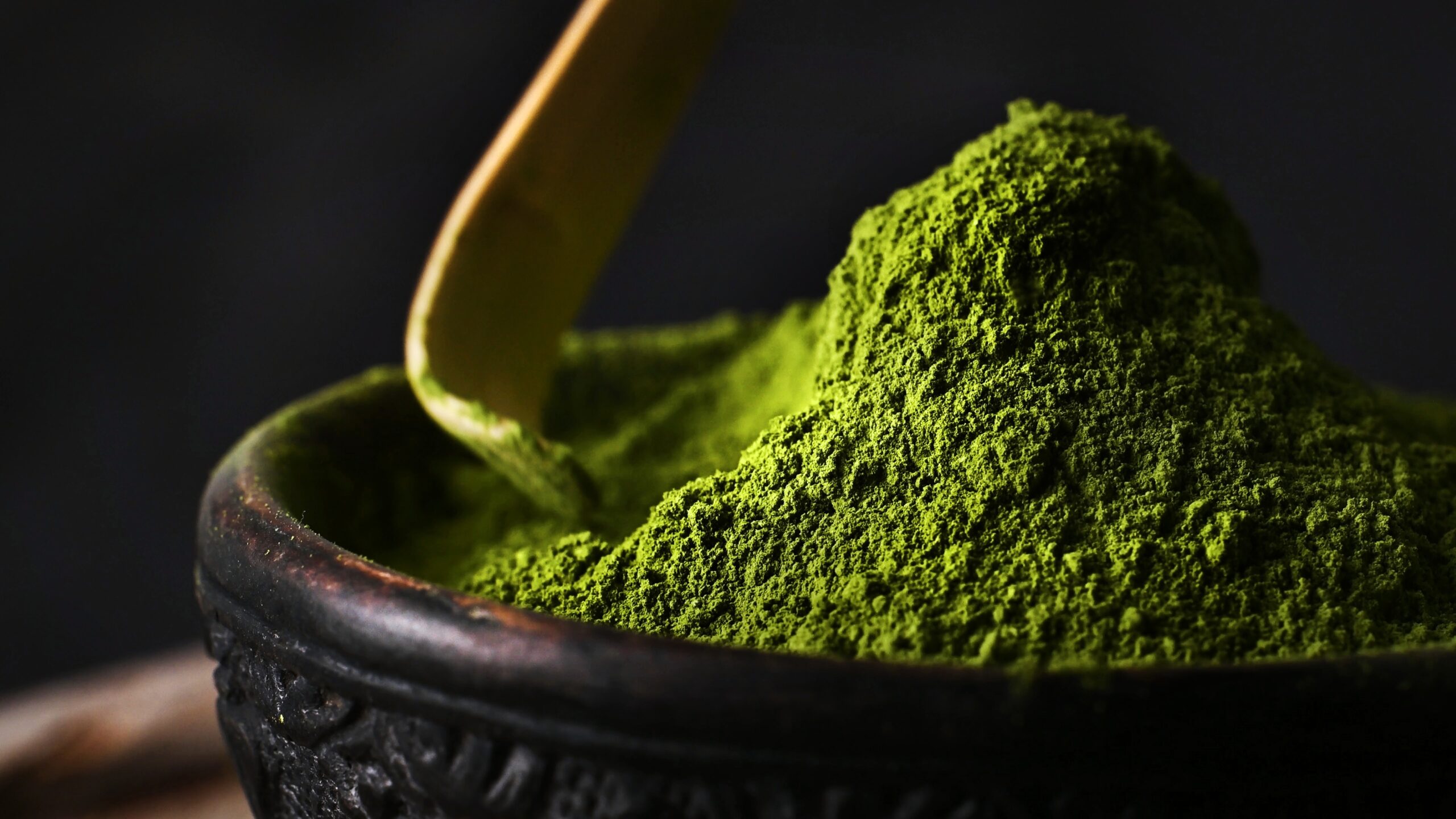 Matcha in an ornate wooden bowl being scooped by a wooden scoop.