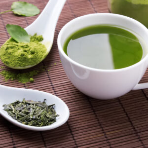 Green tea in a mug with green tea leaves in a spoon and green tea powder in another spoon on a brown, woven mat.