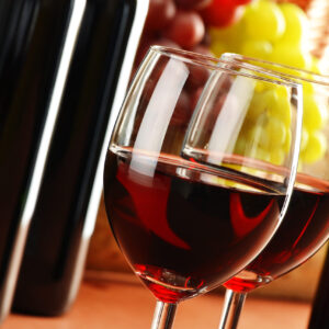 Two full red wineglasses with grapes and wine bottles in the background